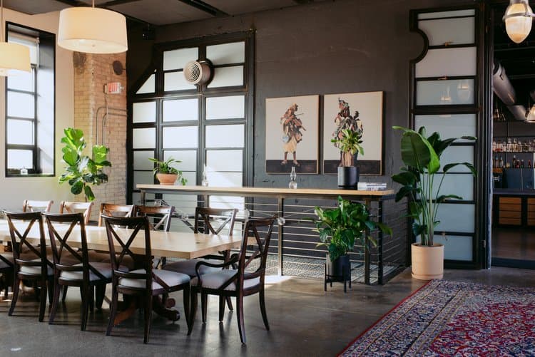 Bright industrial-chic dining space featuring a long wooden table, cross-back chairs, exposed brick walls, and lush green plants.